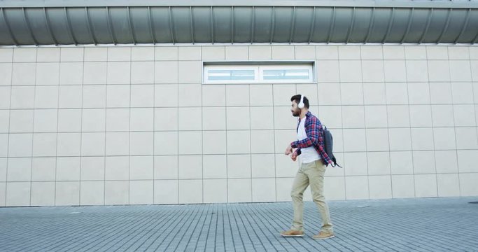 View from the side on the happy cheerful male student with a backpack listening to the music in headphones and moving in the rhythm while walking the street.