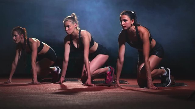 Three sports girls in black clothes of the athlete at night on the treadmill will start for the race at the sprint distance from the sitting position.