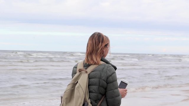 Young woman walking a beach with the phone in hands and taking picture of the sea.
