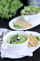 soup puree with green peas and asparagus. decorated with fresh flowers violets. on a black background. in white dishes. bread and rusks in a serving,