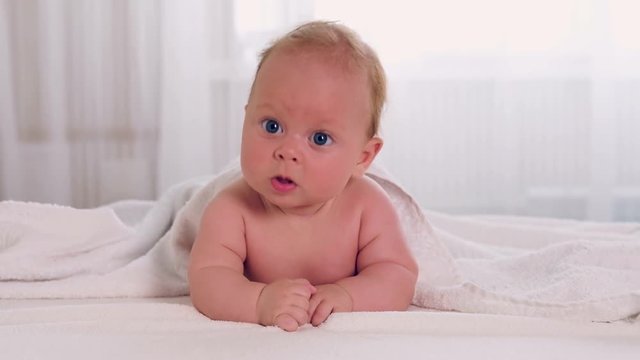 Angelic newborn with beautiful blue eyes lying on belly on the bed and looking aside.