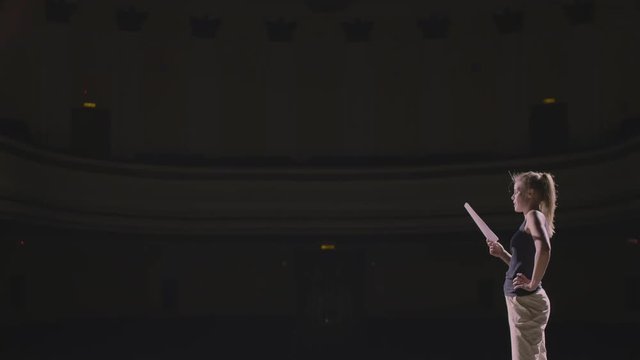 Adult girl walking on the theatre stage and do rehearsal before the premiere. At background empty hall with many rows of seats. The actress concentrated on the script and ready her self for acting