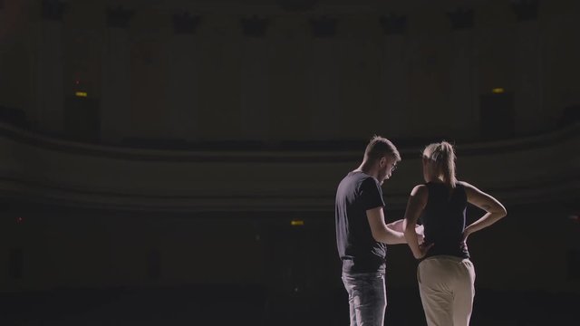 Rehearsal at theatre. Man explain to student actress how to play on the stage on the new performance for public. Dark hall at background. Girl carefully listen the director and say something by answer