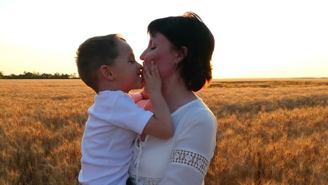 A happy mother holds a child in her arms in a field of wheat against a sunset background. The child kisses the mother's face, the mother kisses the child