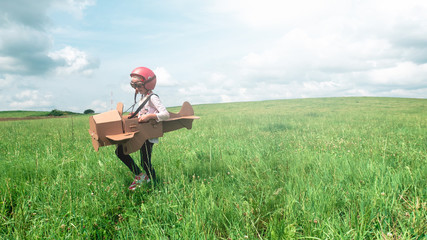 Cute little dreamer kid girl wearing pink helmet and aviator glasses flying in a cardboard airplane through the field, pretending to be a pilot