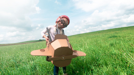 Cute little dreamer kid girl wearing pink helmet and aviator glasses flying in a cardboard airplane through the field, pretending to be a pilot