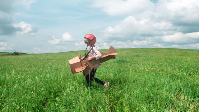 Cute Little Dreamer Kid Girl Wearing Pink Helmet And Aviator Glasses Flying In A Cardboard Airplane Through The Field, Pretending To Be A Pilot