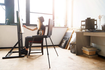 Tattoed amazing blonde curly girl in black hat drawing sketches behind the easel in studio near the sunny window