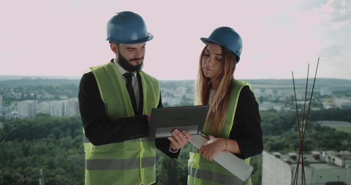 Two young engineers man and female have a discussion on the top of construction site , using a tablet analyzing the plan of building.