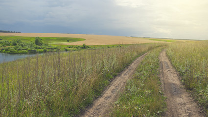 Sunny summer landscape with dirt country road, fields, meadows, woods and pastures.Dark stormy clouds in dramatic overcast sky.Rainy season