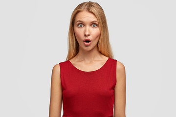Fototapeta premium Astonished young blonde young female model with long hair, keeps jaw dropped, reacts on unexpected news, dressed in red blouse, isolated over white background. Surprised woman stands indoor.