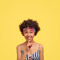 Vertical shot of pleasant looking dark skinned female has positive expression, touches lips with index finger, makes hush gesture, looks upwards as sees something secret, stands against yellow wall