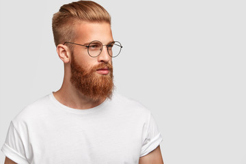 Profile shot of brutal male with thick foxy beard, wears round glasses and looks thoughtfully...