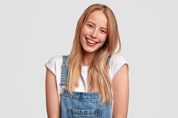 Beautiful happy Caucasian female with charming smile, dressed in fashionable overalls, has long...