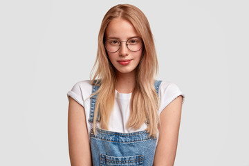 Horizontal portrait of pleasant looking female student in eyewear and denim overalls, ready to listen lectures, prepares for classes, stands against white background. People, youth and style concept