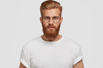 Fototapeta premium Studio shot of confident attractive man with trendy haairstyle and ginger beard, looks mysteriously at camera, wears white t shirt. Handsome unshaven young male freelancer thinks about future
