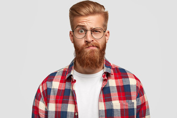 Obraz premium Photo of bewildered puzzled man with thick ginger beard and mustache, raises eyebrows, looks doubtfully at camera, wears fashionable clothes, isolated over white background. Facial expressions