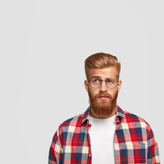 Thoughtful puzzled male student has thick ginger beard looks with hesitation upwards, doesn`t know...
