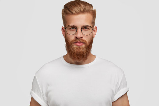 Studio shot of confident attractive man with trendy haairstyle and ginger beard, looks mysteriously at camera, wears white t shirt. Handsome unshaven young male freelancer thinks about future