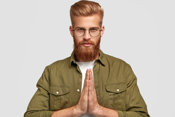 People and religion concept. Serious unshaven young hipster keeps hands in praying gesture,...