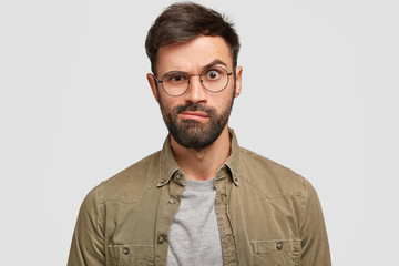 Obraz premium Displeased angry European man raises eyebrows and purses lips furiously, dressed in fashionable shirt, expresses negative emotions, isolated over white background. Facial expressions concept