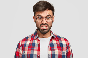 Obraz premium Photo of irritated unshaven man looks angrily at camera, clenches teeth and raises eyebrows, being annoyed with much duties on work, dressed in checkered shirt, stands against white background.