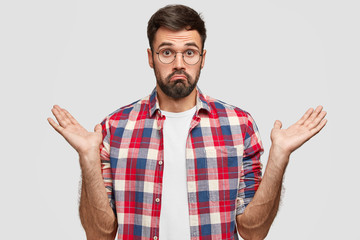 Fototapeta premium Photo of hesitant unshaven male clasps hands with hesitation, has clueless expression, doubts what to do, dressed in checkered shirt, stands against white background. People and confusement concept