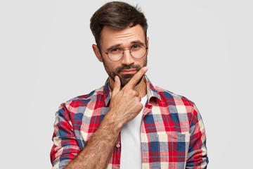 Indoor shot of thoughtful bearded young European man keeps hand under chin, looks seriously at...