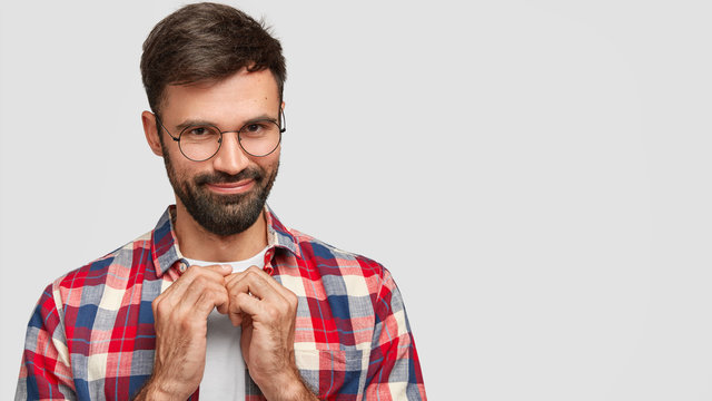 Pleased attractive young bearded man with dark hair, keeps hands together, has intriguing mysterious look, intents prepare surprise to girlfriend, poses over white background with free space
