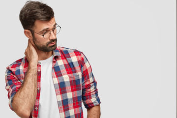 Photo of concentrated thoughtful Caucasian male holds hand on neck, looks curiously aside, notices something interesting, wears checkered casual shirt, isolated over white background with free space