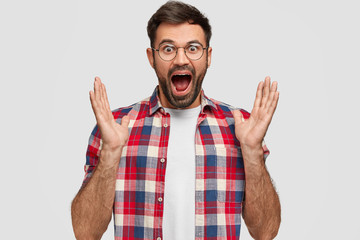 Amazed unshaven young man clasps hands, opens mouth widely, notices something wonderful in front,...