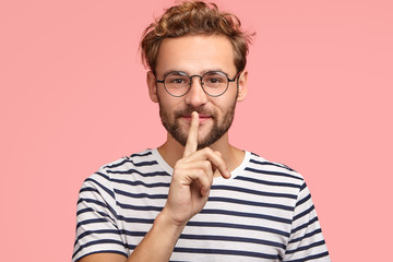 Let`s keep it in secret! Handsome hipster makes shh gesture as spreads rumors, shows hush sign with index finger, dressed casually, isolated over pink background. People, secrecy, conspiracy concept