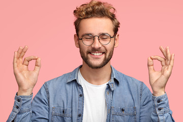 Portrait of cheerful satisfied male with stubble, stands in mudra sign, keeps eyes shut, has...
