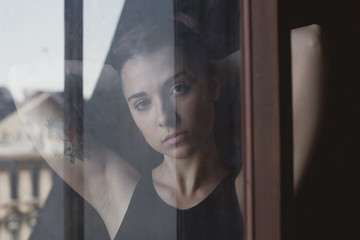 Sensual authentic portrait of young serious lady looking through dirty window, communicating with camera. Real life and art concept. Human feelings, emotions and expressions in natural conditions.