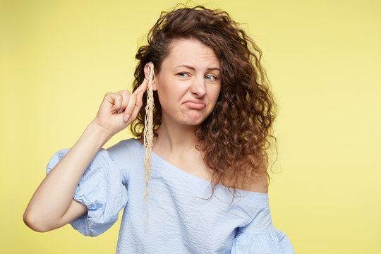 Do not pull my leg. Young beautiful Caucasian female wearing blue blouse, having curly bushy hairstyle and noodles on ear, listening to parter and not believe, feeling cheated,making grimace.