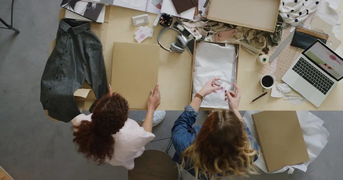 young fashion designer women entrepreneurs packaging clothes in boxes ready for courier shipment colleagues high five celebrating successful sale top view