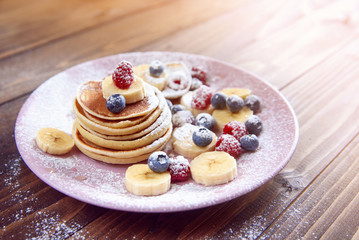 Homemade delicious pancakes with fresh berries sprinkled with powdered sugar in a violet plate on wooden background. A tasty and healthy breakfast of pancakes with raspberries and blueberries.