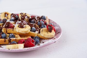 Tasty belgian waffles with blueberries, raspberries, bananas and sugar powder are filled with liquid chocolate.
