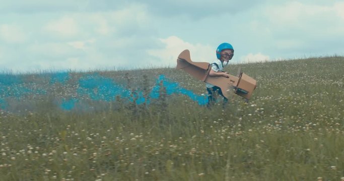 TRACKING Cute little dreamer kid boy wearing blue helmet and aviator glasses flying in a cardboard airplane with attached smoke bombs through the field, pretending to be a pilot. 4K UHD 60 FPS SLO MO 
