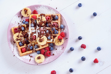 Tasty belgian waffles with blueberries, raspberries, bananas and sugar powder are filled with liquid chocolate. Top view.