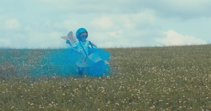 TRACKING Cute little dreamer kid boy wearing blue helmet and aviator glasses flying in a cardboard airplane with attached smoke bombs through the field, pretending to be a pilot. 4K UHD 60 FPS SLO MO 