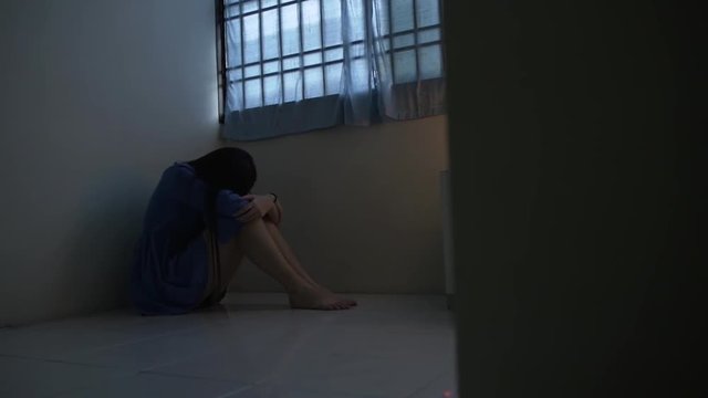A sad young girl sits in the corner of an empty room with bars on the window. Tracking shot in slow motion.