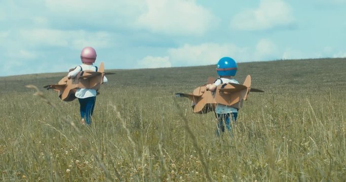 TRACKING Cute little dreamer siblings boy and girl wearing helmets and aviator glasses flying in a cardboard airplanes through the field, pretending to be pilots. 4K UHD 60 FPS SLO MO 