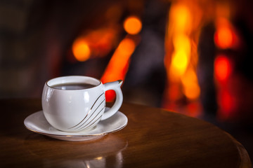 White cup of tea or coffee near fireplace on wooden table. Winter and Christmas holiday concept