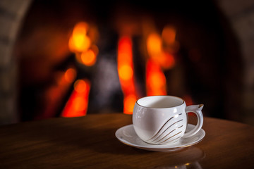 White cup of tea or coffee near fireplace on wooden table. Winter and Christmas holiday concept