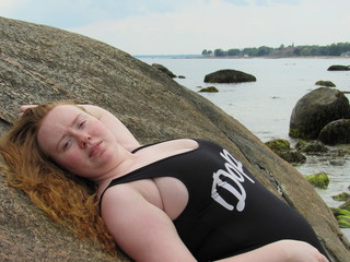 A beautiful young redheaded woman leaning against a rock sunbathing on the beach with a black one-piece bathing suit that says 