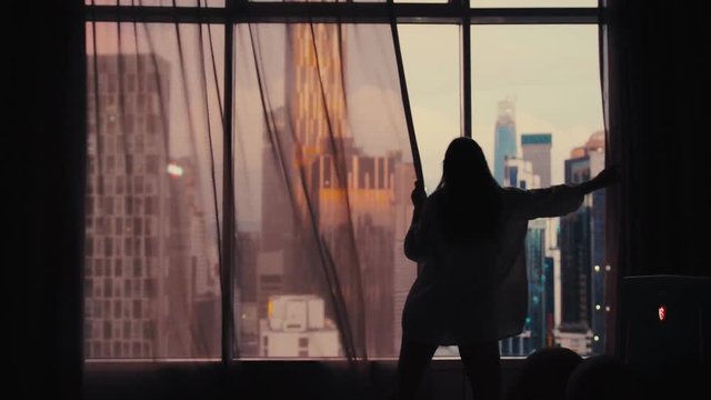 Young woman uncover the big window in apartment on the city buildings background. Sunset in the city. slow motion. 3840x2160