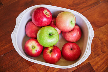 Top view of red apples and green apple in the white plastic bowl on brown wooden floor