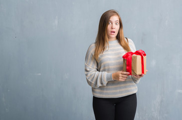 Young adult woman over grey grunge wall holding a present scared in shock with a surprise face, afraid and excited with fear expression