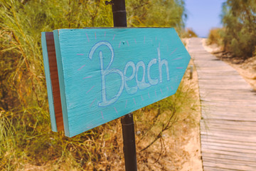 hand written wooden blue, turquoise sign on a metal pole  pointing to the beach which is reached along a wooden path walkway